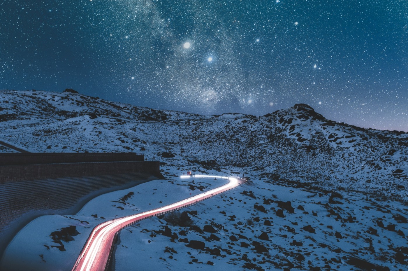 A trail of light follows a winding road through snow-covered mountains beneath a star-filled night sky.